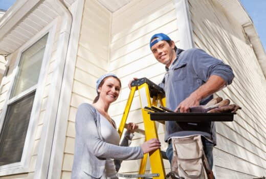 Couple doing home repairs outside.  Man (30s) standing on ladder with toolbox.